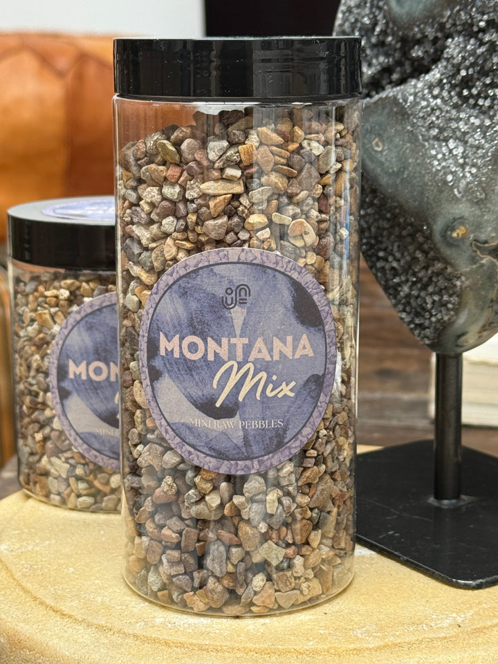 A clear cylindrical jar filled with small decorative stones labeled 'Montana Mix' sits on a wooden surface, with a second matching jar behind it and a dark textured object at the right edge.