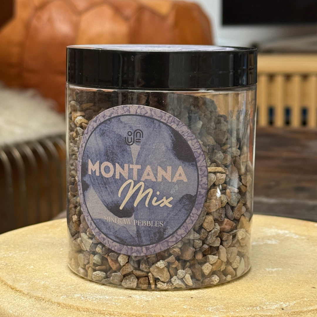 A clear plastic jar filled with Montana Mix decorative pebbles, featuring a black lid and a round purple label, set on a wooden surface with a blurred indoor background.